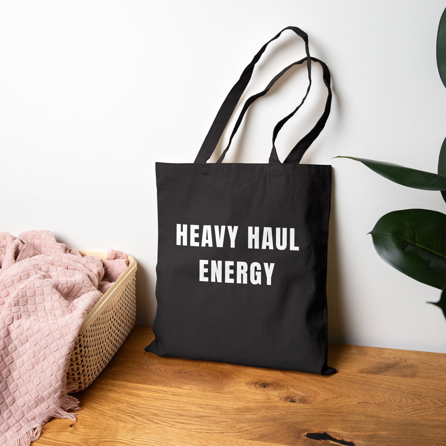 Black tote bag with 'HEAVY HAUL ENERGY' text on a wooden surface with a plant in the background