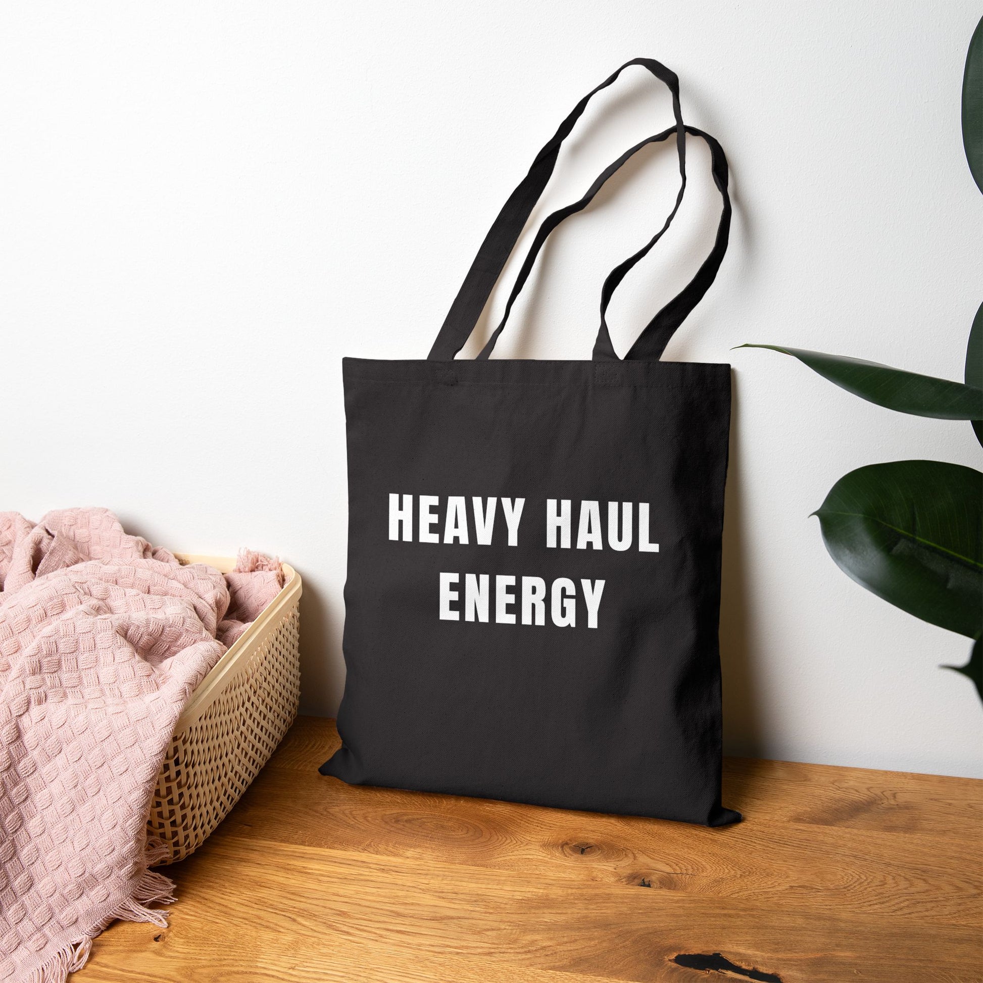 Black tote bag with 'HEAVY HAUL ENERGY' text on a wooden surface with a plant in the background