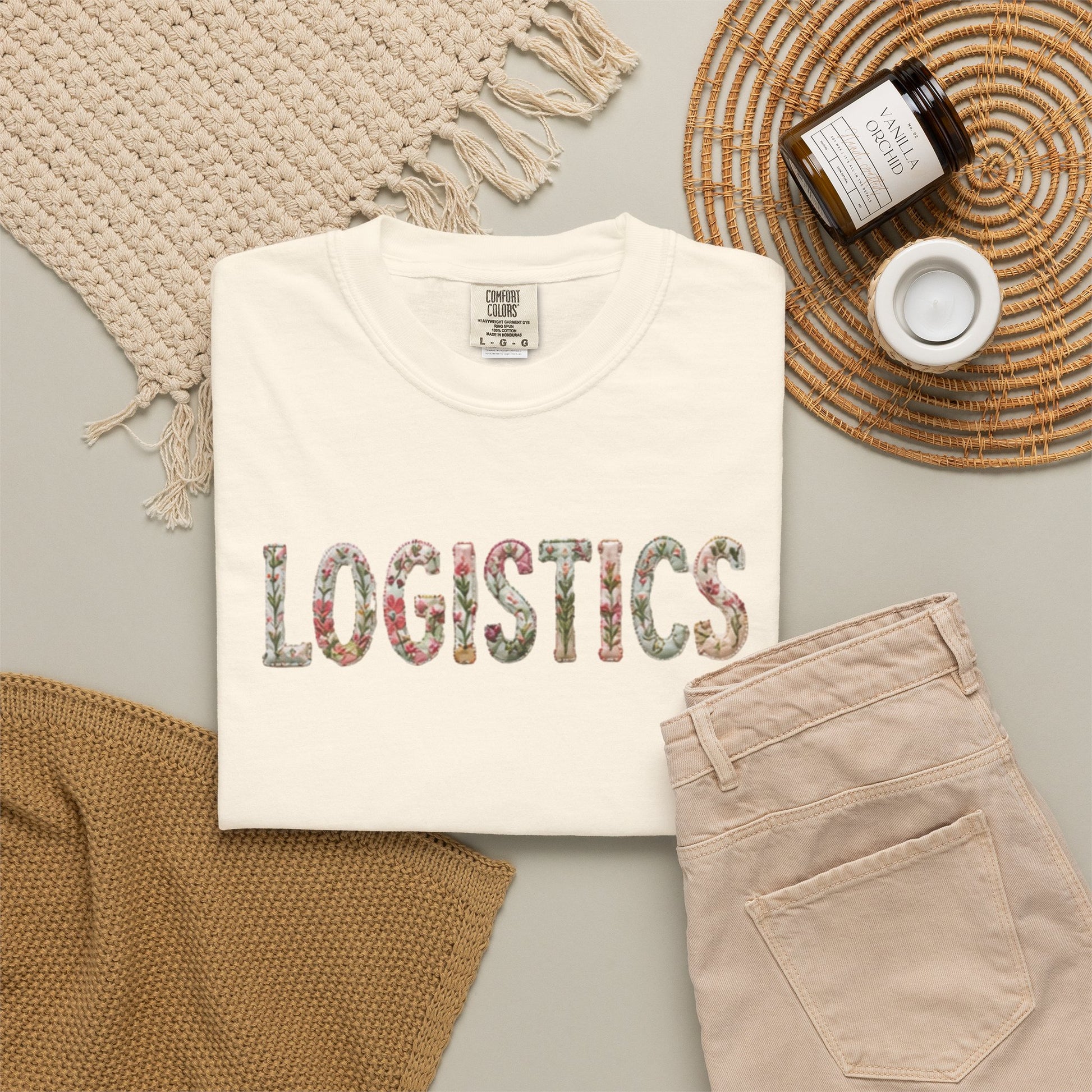 White t-shirt with 'LOGISTICS' printed on it, placed on a surface with beige pants and decorative items.