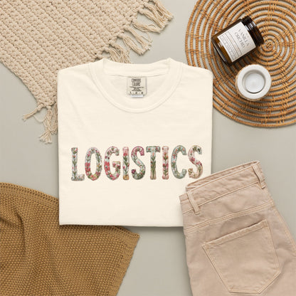 White t-shirt with 'LOGISTICS' printed on it, placed on a surface with beige pants and decorative items.