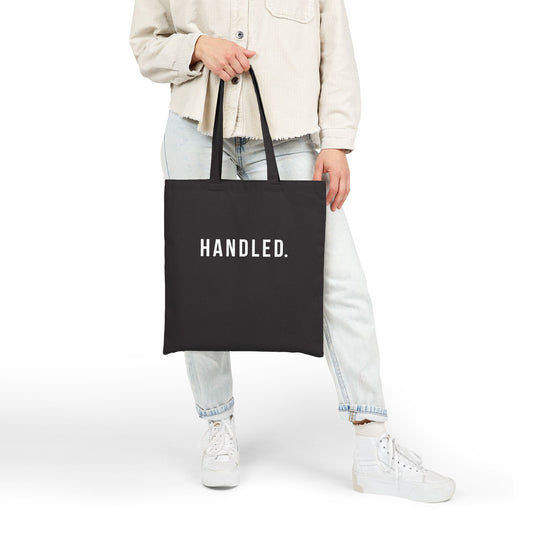 Person holding a black tote bag with 'HANDLED.' text on a white background