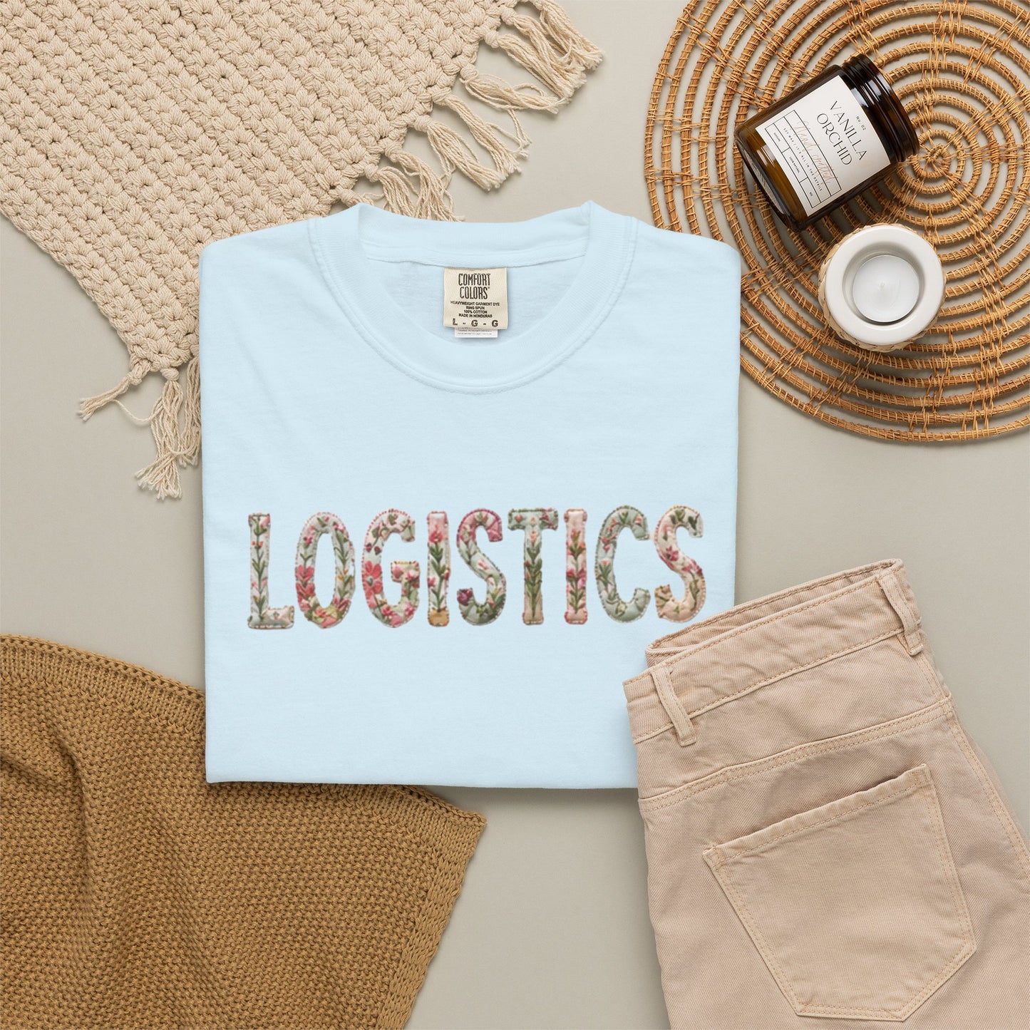 Light blue t-shirt with 'LOGISTICS' printed on it, placed on a textured surface with a bottle and a container.