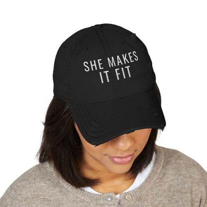 Black cap with 'SHE MAKES IT FIT' text worn by a person on a white background