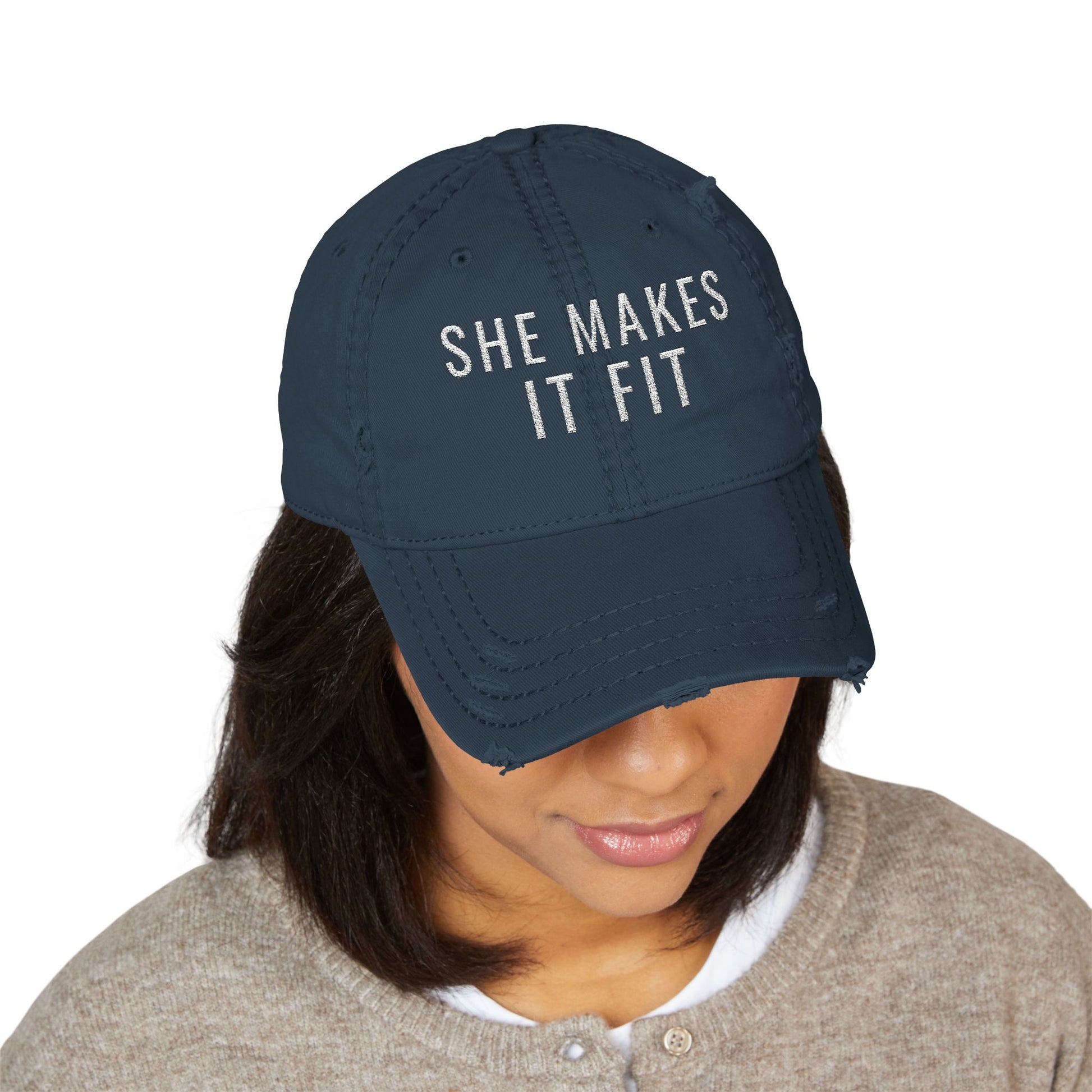 Navy blue cap with 'SHE MAKES IT FIT' text worn by a person on a white background