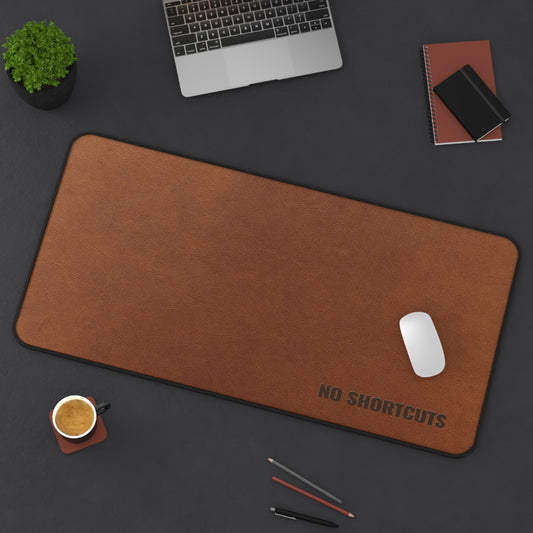 Brown desk mat with 'NO SHORTCUTS' text on a dark surface with office items.