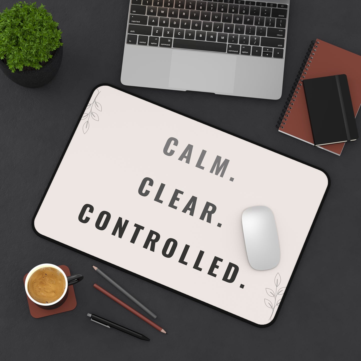Desk setup with a mouse pad featuring 'CALM. CLEAR. CONTROLLED.' text, laptop, notebook, and coffee cup.