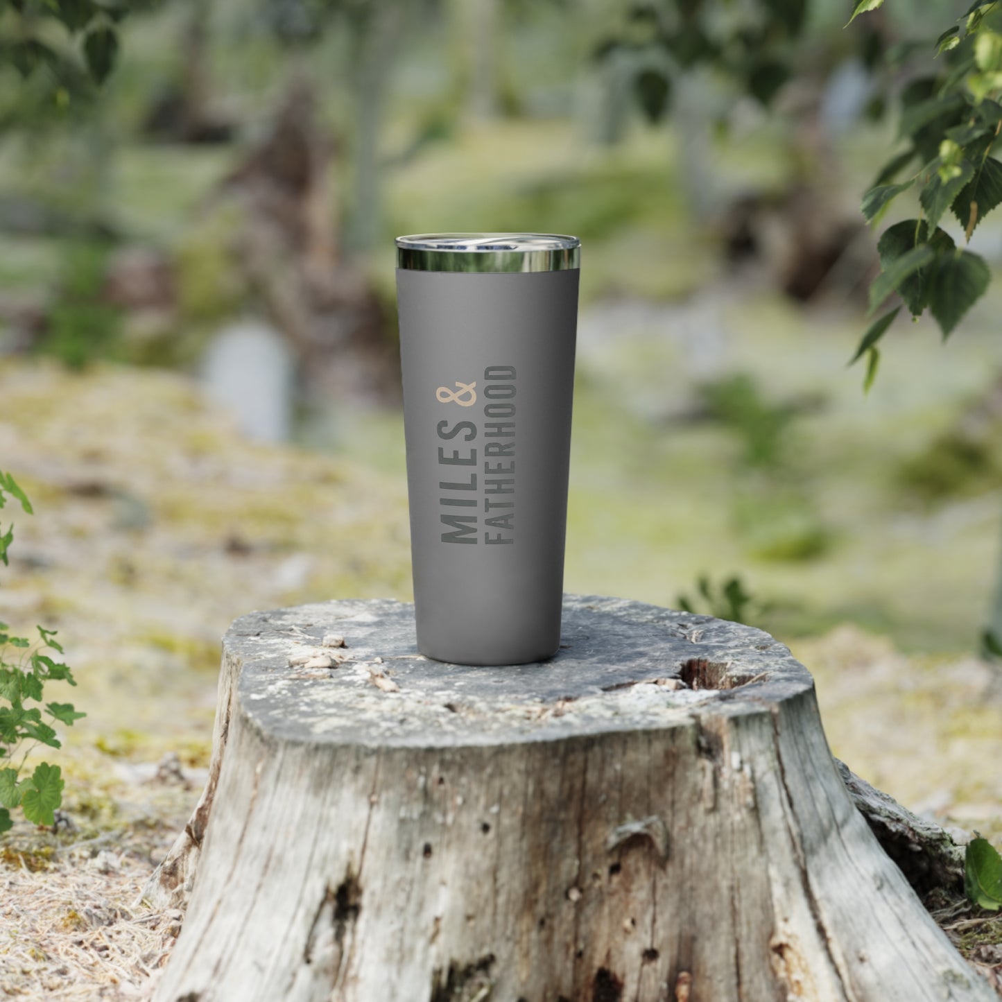 Gray tumbler with 'MILES & FATHERHOOD' text on a wooden stump in a natural setting