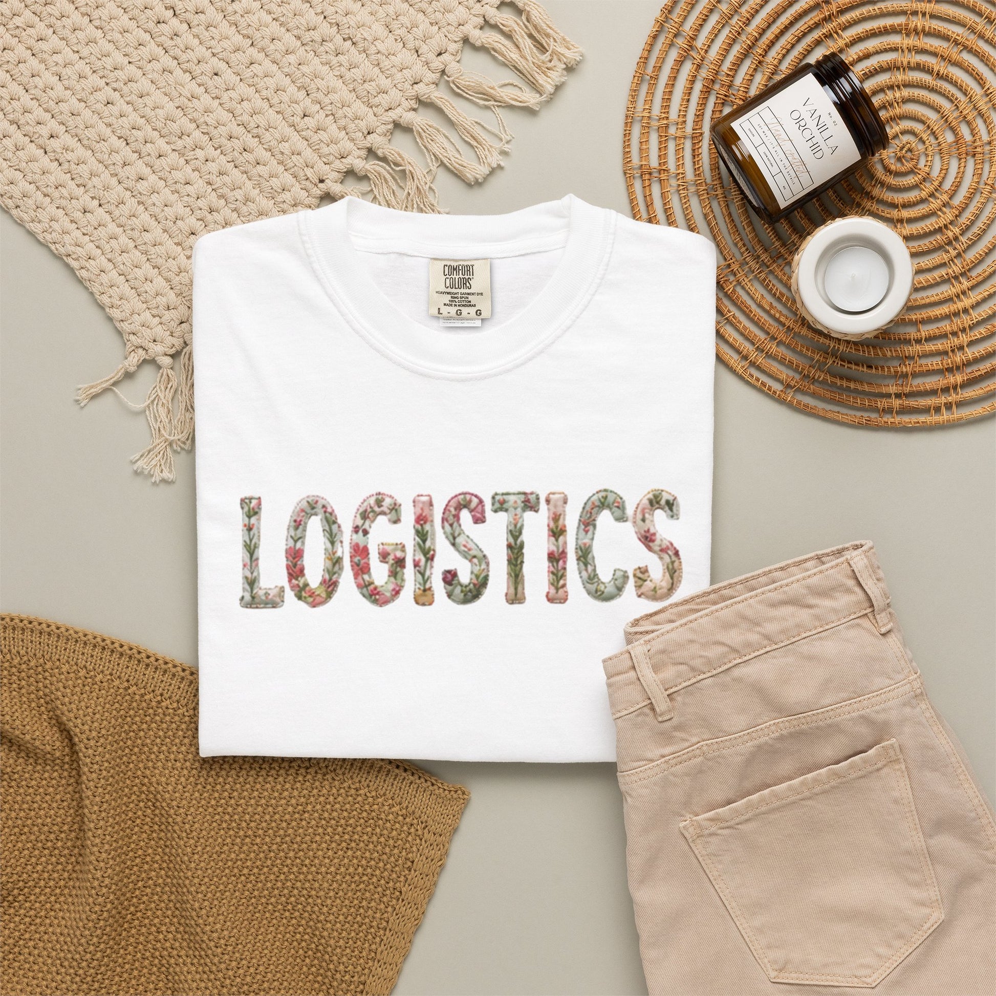 White t-shirt with 'LOGISTICS' printed on it, placed on a surface with beige pants and decorative items.