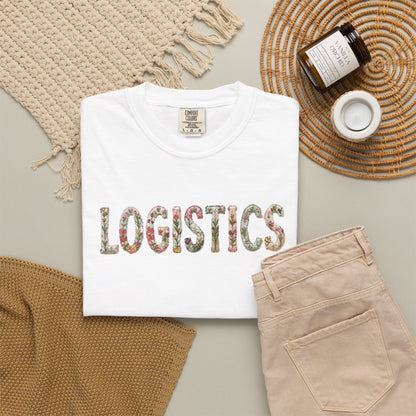 White t-shirt with 'LOGISTICS' printed on it, placed on a surface with beige pants and decorative items.