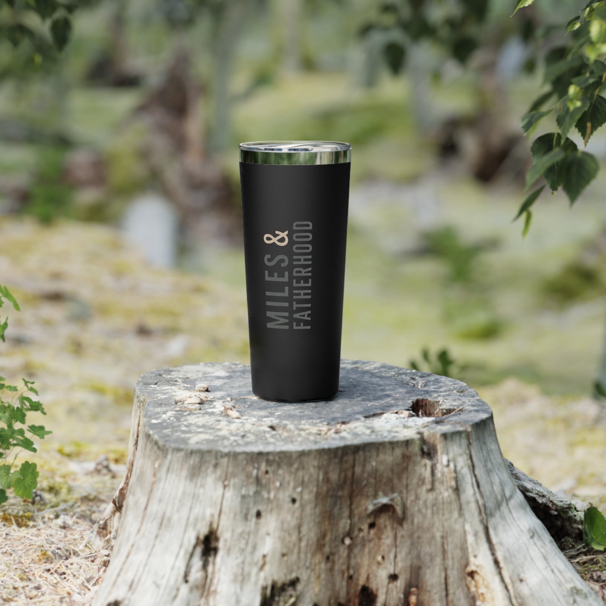 Black tumbler with 'Miles & Fatherhood' text on a wooden stump outdoors