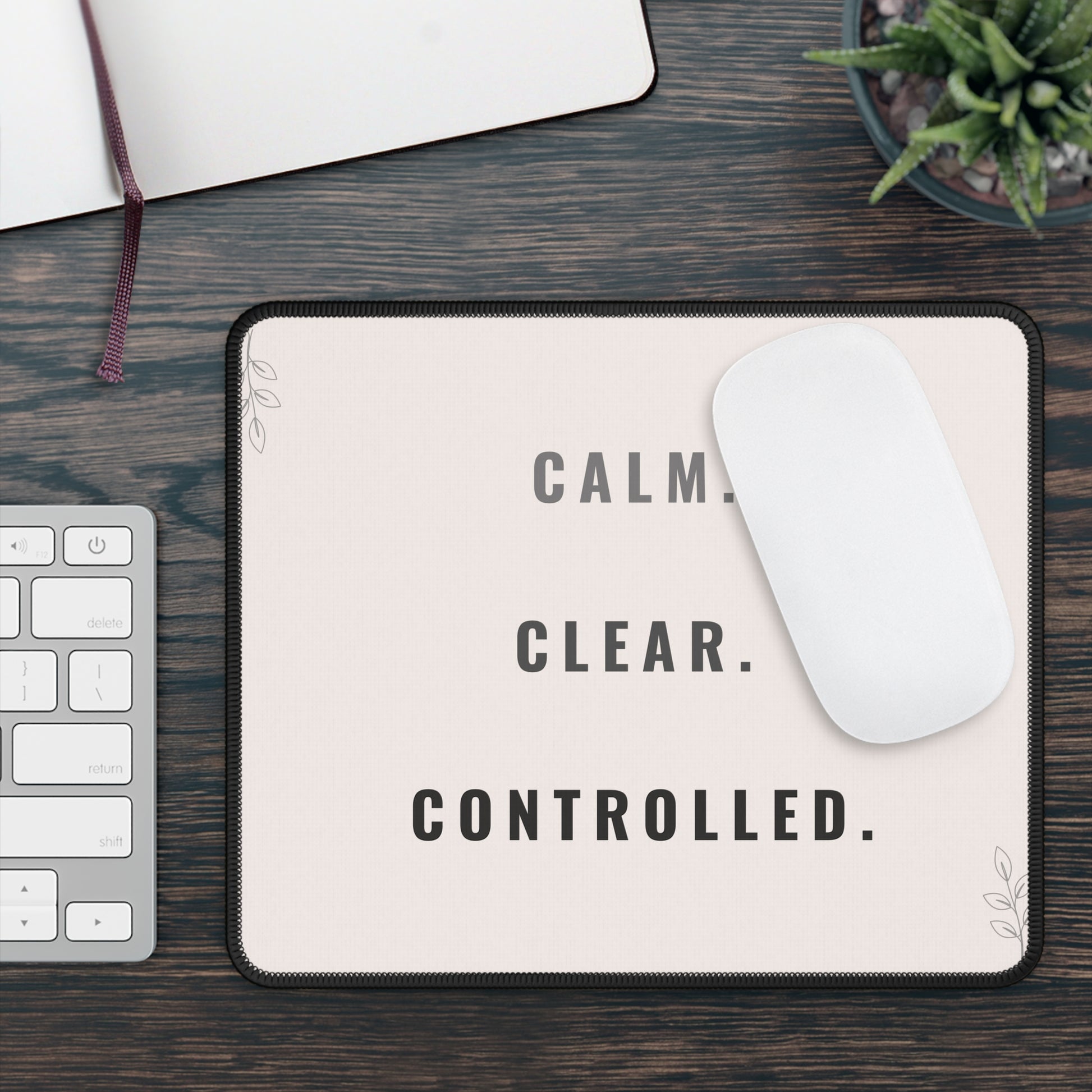 Mouse pad with 'CALM. CLEAR. CONTROLLED.' text on a desk with keyboard and plant