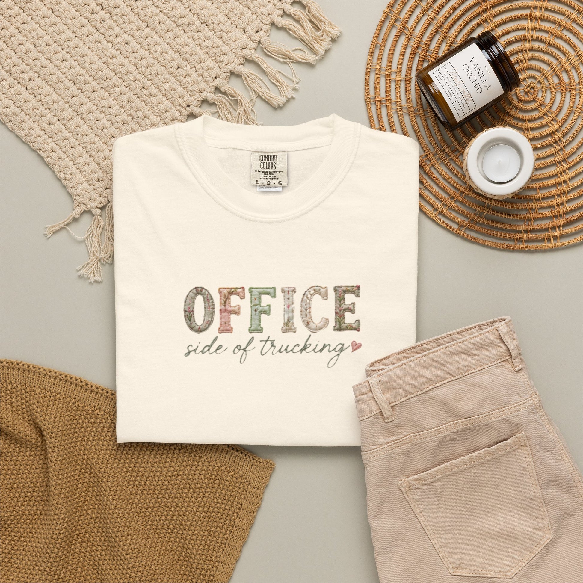 White t-shirt with 'OFFICE SIDE OF TRUCKING' text, beige pants, and decorative items on a light gray surface.