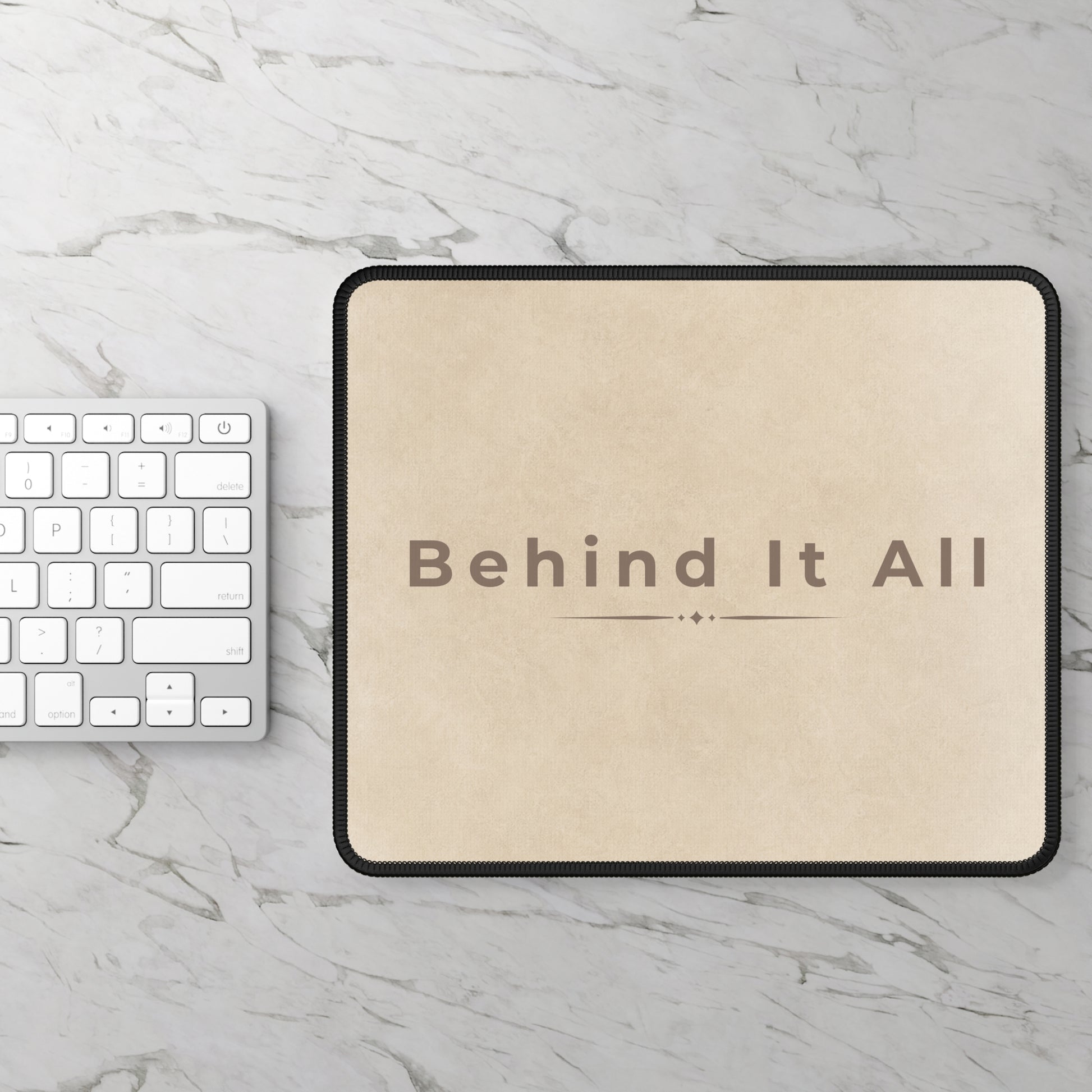 Beige mouse pad with 'Behind It All' text on a marble surface with a keyboard.