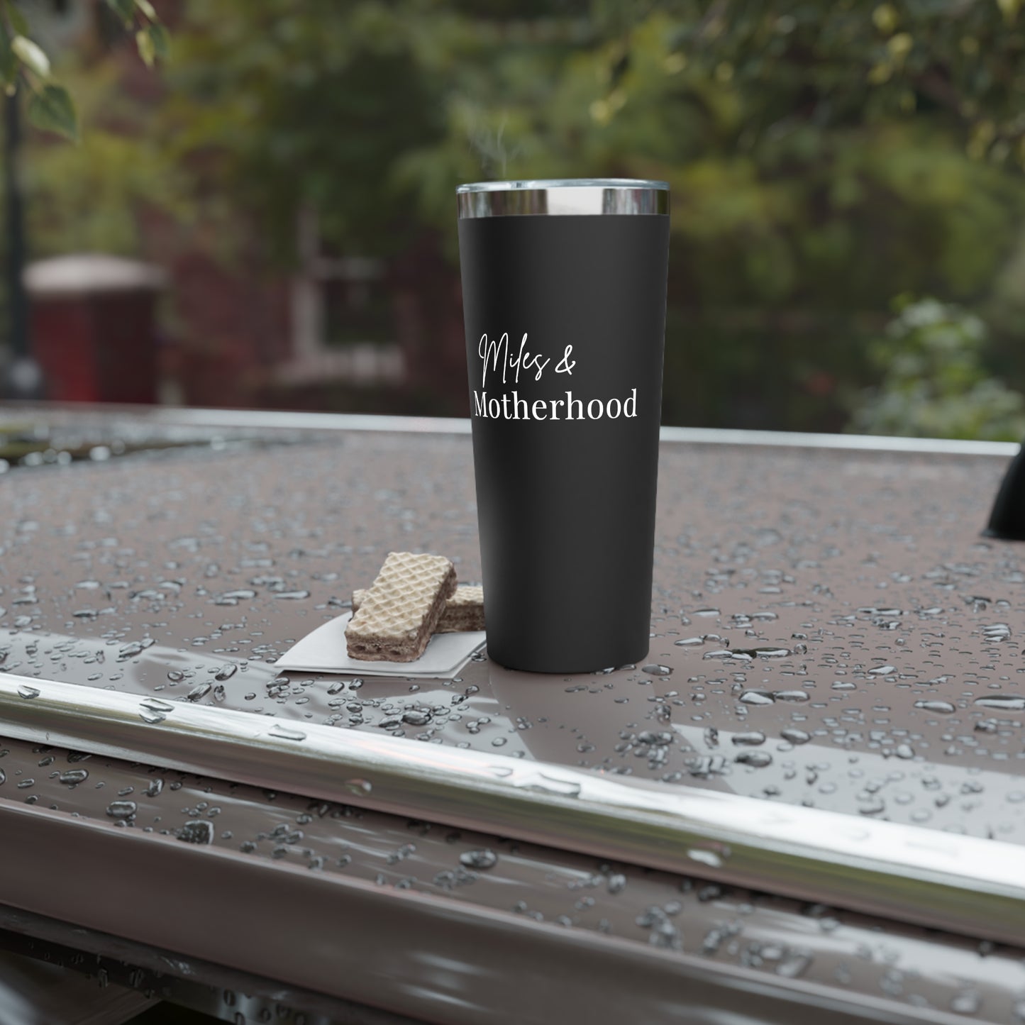 Black tumbler with 'Miles & Motherhood' text on a wet surface outdoors