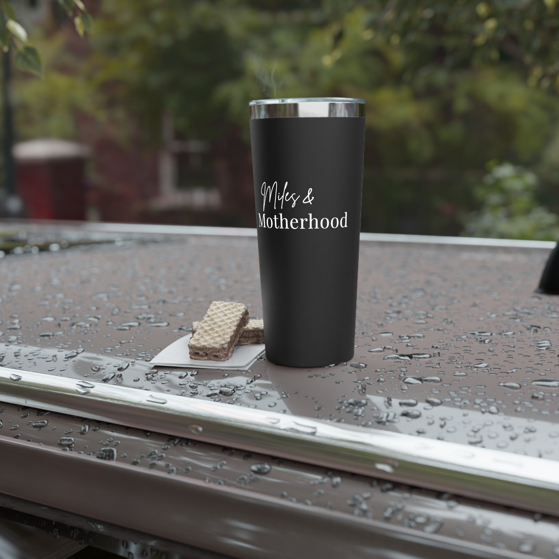 Black tumbler with 'Miles & Motherhood' text on a wet surface outdoors
