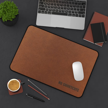 Brown mouse pad with 'NO SHORTCUTS' text on a dark surface with laptop, mouse, and office supplies.
