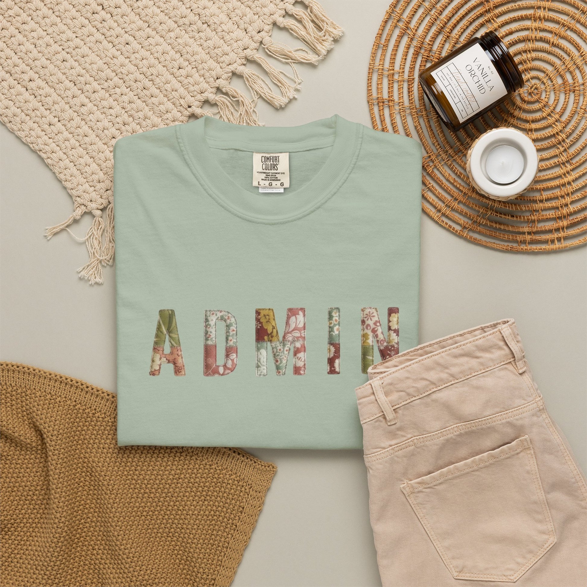 Folded mint green shirt with 'ADMIN' printed in colorful letters on a neutral surface with a woven mat and beige pants.