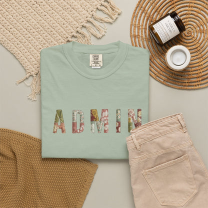 Folded mint green shirt with 'ADMIN' printed in colorful letters on a neutral surface with a woven mat and beige pants.