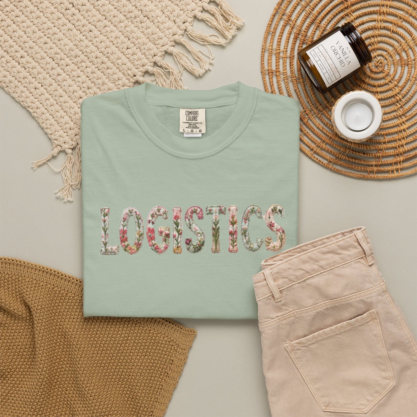 Folded mint green sweatshirt with floral 'LOGISTICS' text on a neutral surface with accessories.