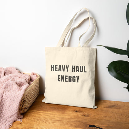 Beige tote bag with 'HEAVY HAUL ENERGY' text on a wooden surface with a plant in the background