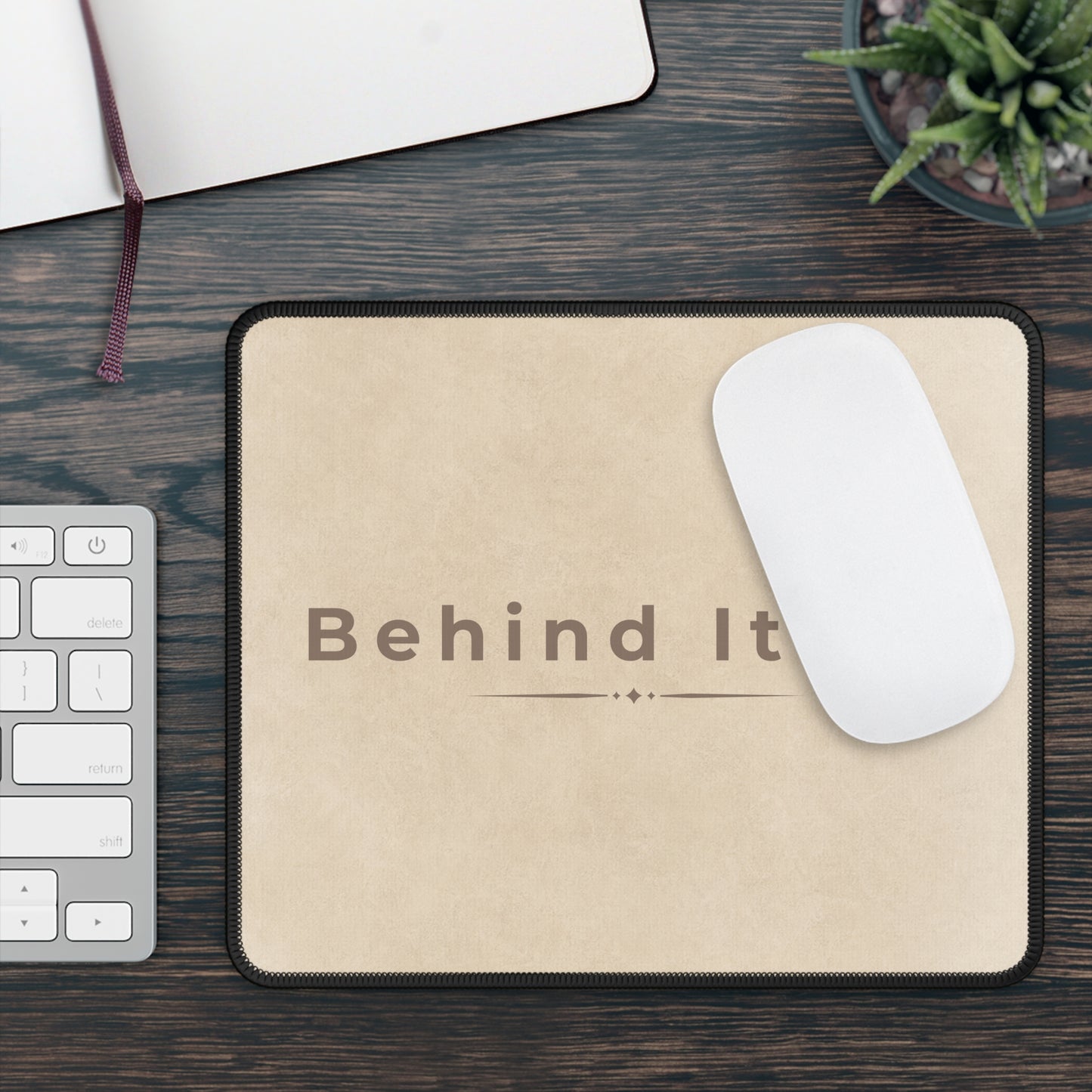 Beige mouse pad with 'Behind It' text on a wooden desk with a keyboard and plant.