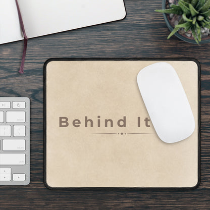 Beige mouse pad with 'Behind It' text on a wooden desk with a keyboard and plant.